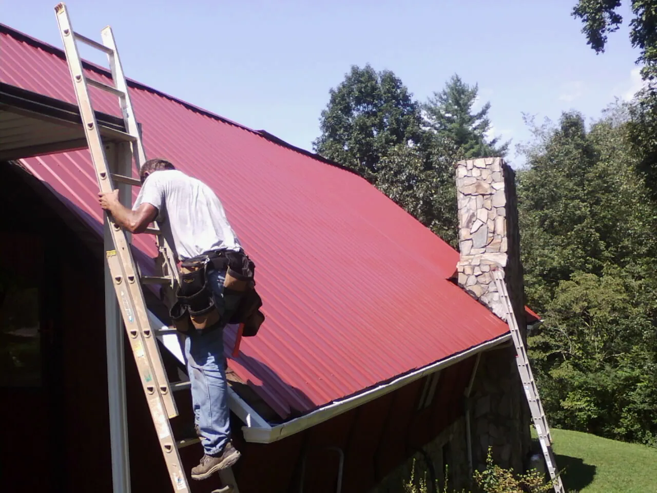 Roofer on a ladder beside a red metal roof and stone chimney — Soffit & Fascia Repair by Roofing Brothers in Lynden